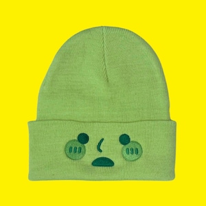 May include: A light green beanie with an embroidered face design. The face features two circular eyes, two cheek circles, a curved nose, and a downturned mouth. The beanie is made of a soft knit material and is set against a bright yellow background.