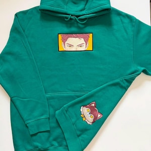 May include: A teal hooded sweatshirt featuring an embroidered rectangular patch on the chest displaying a pair of anime-style eyes with pink hair. A smaller embroidered patch of a stylized brown and white animal head is on the lower sleeve.
