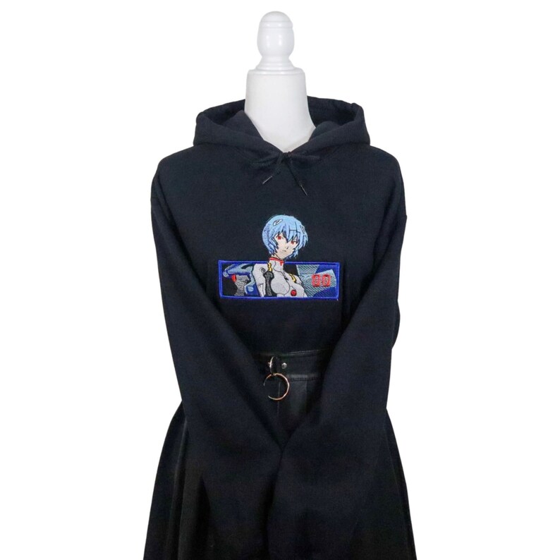 May include: Black hooded sweatshirt with an embroidered image of an anime character with blue hair and a red collar. The character is surrounded by a blue rectangle with the text "00" in red.