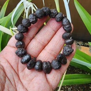 Puede incluir: Una pulsera de cuentas marrón oscuro compuesta por pequeñas cuentas redondas de forma irregular. La pulsera se sostiene en una mano, con un fondo borroso de hojas verdes y una flor blanca.