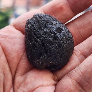 Pode incluir: Uma rocha escura de forma irregular com uma superfície áspera e texturizada. A rocha é segurada em uma mão, mostrando sua cor escura e sua forma única. O objeto parece ser uma pedra natural ou um espécime mineral.