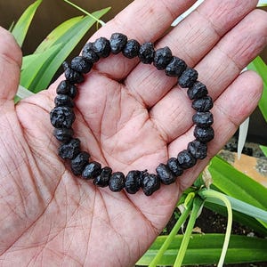 Puede incluir: Un brazalete de cuentas negras compuesto por cuentas de forma irregular. El brazalete se muestra en una mano, con follaje verde y un fondo marrón. Las cuentas parecen estar hechas de un material oscuro, posiblemente natural.