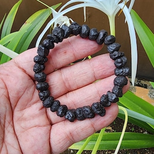 Puede incluir: Un brazalete de cuentas negras compuesto por piedras de forma irregular. El brazalete se sostiene en una mano, con un fondo de hojas verdes y flores blancas. Las piedras miden aproximadamente 1,3 cm de diámetro.