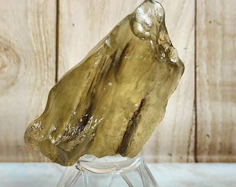 May include: A translucent, irregularly shaped, yellow-brown gemstone. The stone has dark brown inclusions and is displayed on a clear, faceted stand. The background features a light wood grain pattern.