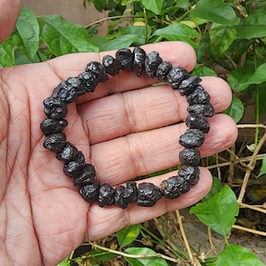 Puede incluir: Un brazalete de cuentas negras compuesto por piedras de forma irregular. El brazalete está ensartado, formando una forma circular. Las piedras tienen una superficie rugosa y texturizada. El brazalete se muestra sobre un fondo de follaje verde.