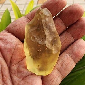 May include: A translucent, pale yellow raw gemstone with an irregular, organic shape. The stone features a smooth, polished-looking surface on one side and a rough, textured, matte-finish section along the top edge, displaying natural variations.