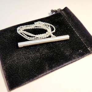 May include: A silver bar pendant necklace on a silver chain, displayed on a black velvet pouch.