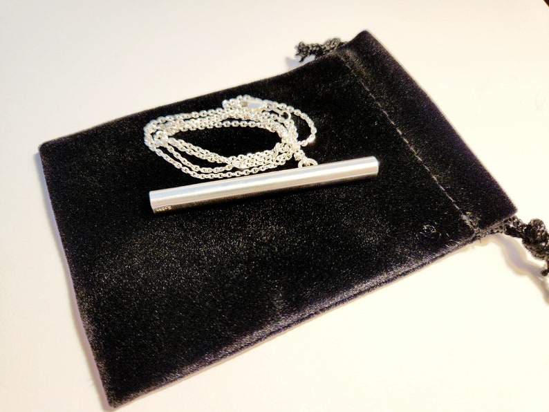 May include: A silver bar pendant necklace on a silver chain, displayed on a black velvet pouch.