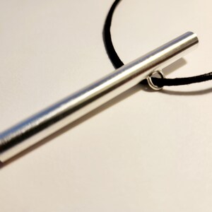 May include: A silver metal bar pendant on a black cord necklace. The bar is smooth and has a slightly rounded end.