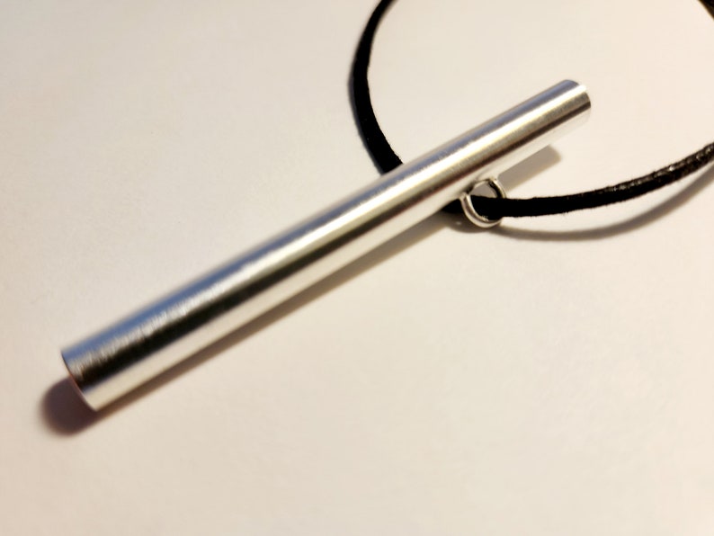 May include: A silver metal bar pendant on a black cord necklace. The bar is smooth and has a slightly rounded end.