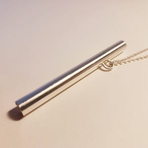 May include: A silver necklace with a long, cylindrical pendant. The pendant is smooth and has a slightly rounded end.