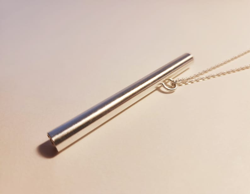 May include: A silver necklace with a long, cylindrical pendant. The pendant is smooth and has a slightly rounded end.