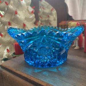 May include: A vibrant blue glass bowl with a starburst pattern on the base and sides. The bowl has a scalloped rim and is sitting on a wooden surface. The bowl is likely vintage or antique.