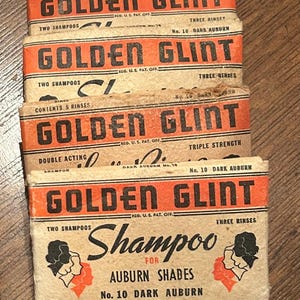 May include: Vintage Golden Glint shampoo boxes in a stack. The boxes are tan with orange and black text, advertising shampoo for auburn shades. The text includes "Brightens Natural Color" and "No. 10 Dark Auburn."