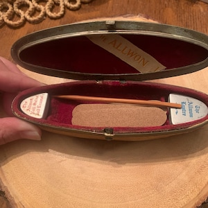 May include: An oval-shaped, silver-colored metal box with a red velvet interior. The box is open and contains a small wooden stick and a small, white, rectangular card with the text "The Allwon Razor" printed on it.
