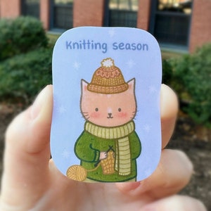 May include: A sticker featuring a cartoon cat knitting. The cat wears a green sweater, a beige scarf, and a matching hat with a white pom-pom. The sticker has the text "Knitting season" on a light blue background with snowflakes.