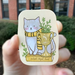 May include: A whimsical illustration of a gray cat wearing a yellow and black scarf, sitting on a book. The cat is next to a golden trophy filled with greenery. The text on the book reads "patient, loyal, kind."