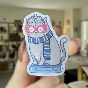 May include: A sticker featuring a gray cat wearing a tiara, pink glasses, and a blue and white striped scarf. The cat sits on a blue book with the words "Wise, Creative, sharp" printed on it. The sticker has a white border.