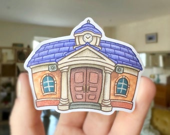 May include: A sticker featuring a cartoon illustration of a building with a blue roof, clock tower, and brown doors. The building has orange walls and blue window accents. The sticker is held in a person's hand.