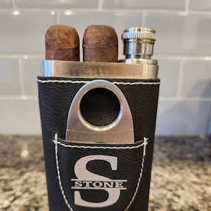 May include: A black leather flask with a built-in cigar cutter and two cigars. The flask has a silver lid and a silver monogram with the letter "S" and the word "STONE" underneath.