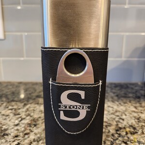 May include: A black leather cigar case with a silver metal top and a built-in cutter. The case has a white stitched border and a white monogram that reads "STONE".