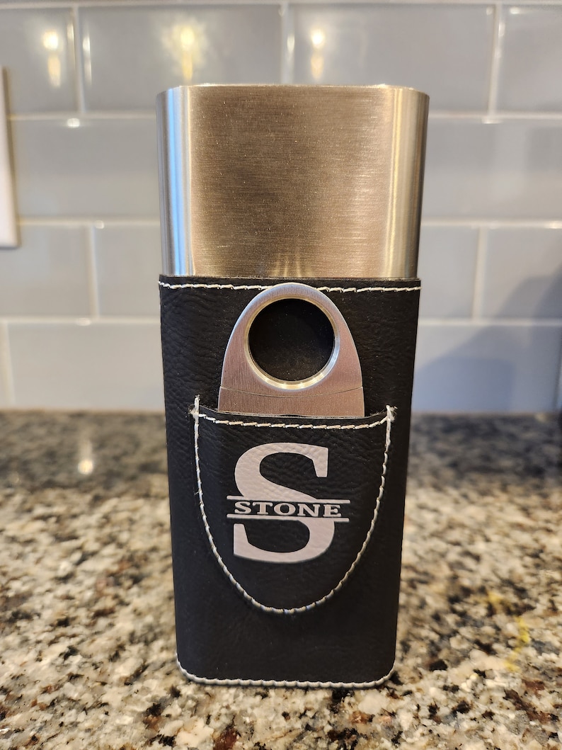 May include: A black leather cigar case with a silver metal top and a built-in cutter. The case has a white stitched border and a white monogram that reads "STONE".