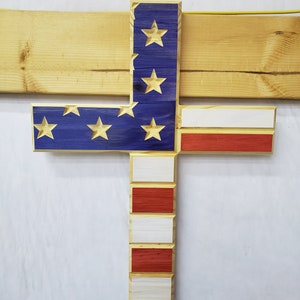 Könnte beinhalten: Ein Holzkreuz, das mit der amerikanischen Flagge bemalt ist. Das Kreuz besteht aus einzelnen Holzbrettern, und die Flagge ist in den traditionellen Farben Rot, Weiß und Blau bemalt. Die Sterne sind weiß und die Streifen sind rot und weiß.