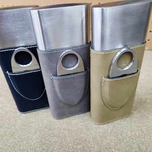 May include: Three silver cigar cases with leather coverings. The cases have a built-in cigar cutter. The cases are in black, grey, and brown.