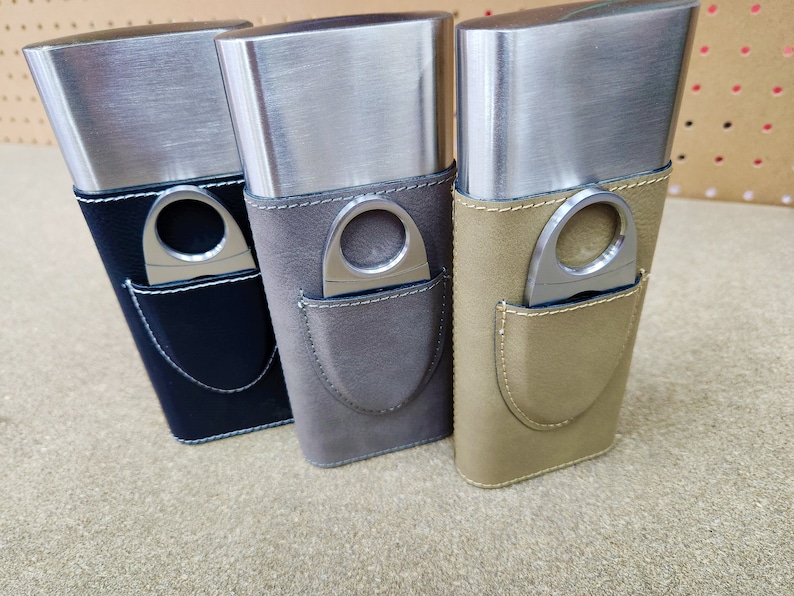 May include: Three silver cigar cases with leather coverings. The cases have a built-in cigar cutter. The cases are in black, grey, and brown.