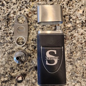May include: A black leather flask with a silver top and a silver monogrammed shield featuring the letter "S" and the word "STONE" below. The flask is accompanied by a silver cigar cutter and a silver funnel.