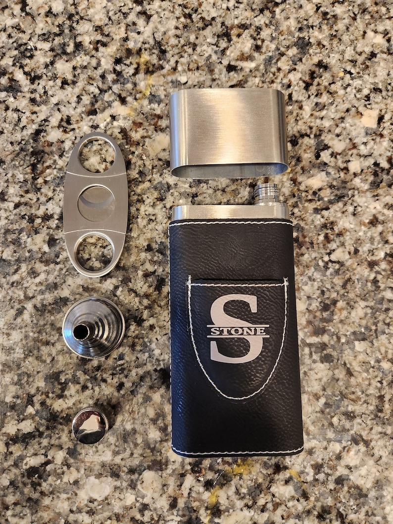 May include: A black leather flask with a silver top and a silver monogrammed shield featuring the letter "S" and the word "STONE" below. The flask is accompanied by a silver cigar cutter and a silver funnel.