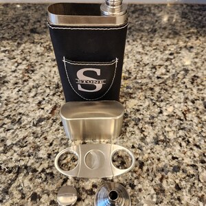 May include: A black leather flask with a silver top and a silver monogrammed patch that says "STONE" with the letter "S" in a larger font. The flask is accompanied by a silver cigar cutter, a silver funnel, and a silver cap.