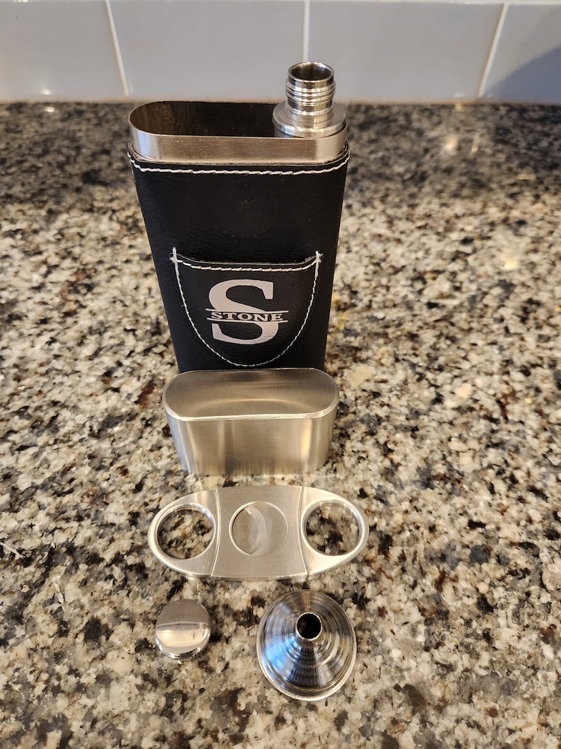 May include: A black leather flask with a silver top and a silver monogrammed patch that says "STONE" with the letter "S" in a larger font. The flask is accompanied by a silver cigar cutter, a silver funnel, and a silver cap.