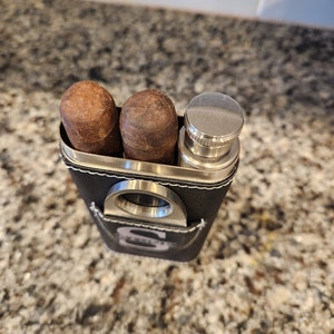 May include: A black leather travel case with a silver cigar cutter, two brown cigars, and a silver flask.