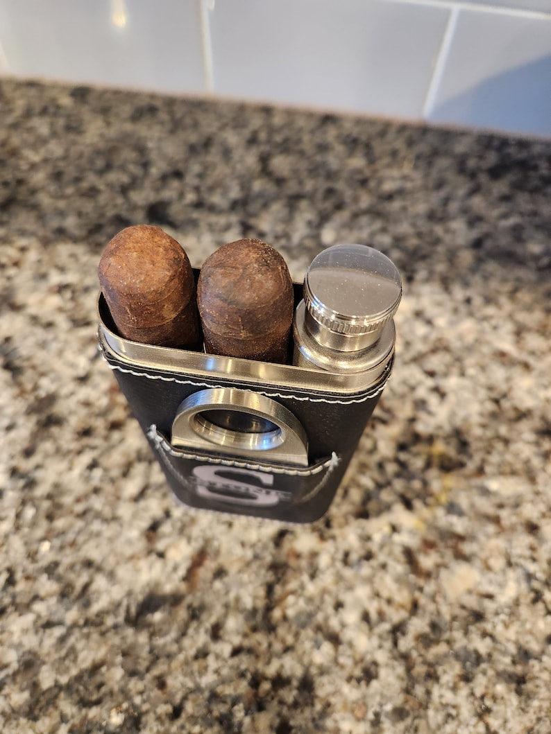 May include: A black leather travel case with a silver cigar cutter, two brown cigars, and a silver flask.
