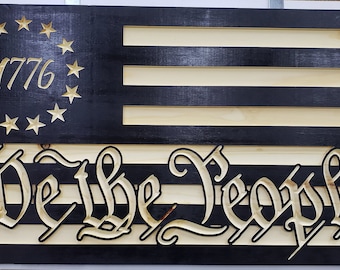 May include: A black and white wooden American flag with 13 stripes and a circle of 13 stars with the year 1776 inside. The words "We the People" are carved into the bottom of the flag.