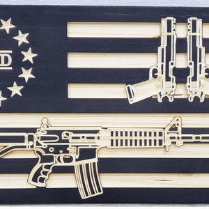 May include: A wooden American flag with a black background and natural wood stripes. The flag features a stylized image of a rifle and two handguns, along with the text "2ND" in a circle of stars.