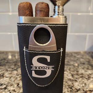 May include: A black leather cigar case with a silver metal cigar cutter, a silver metal funnel, and two cigars. The case has a pocket with a silver metal monogram that says "STONE".