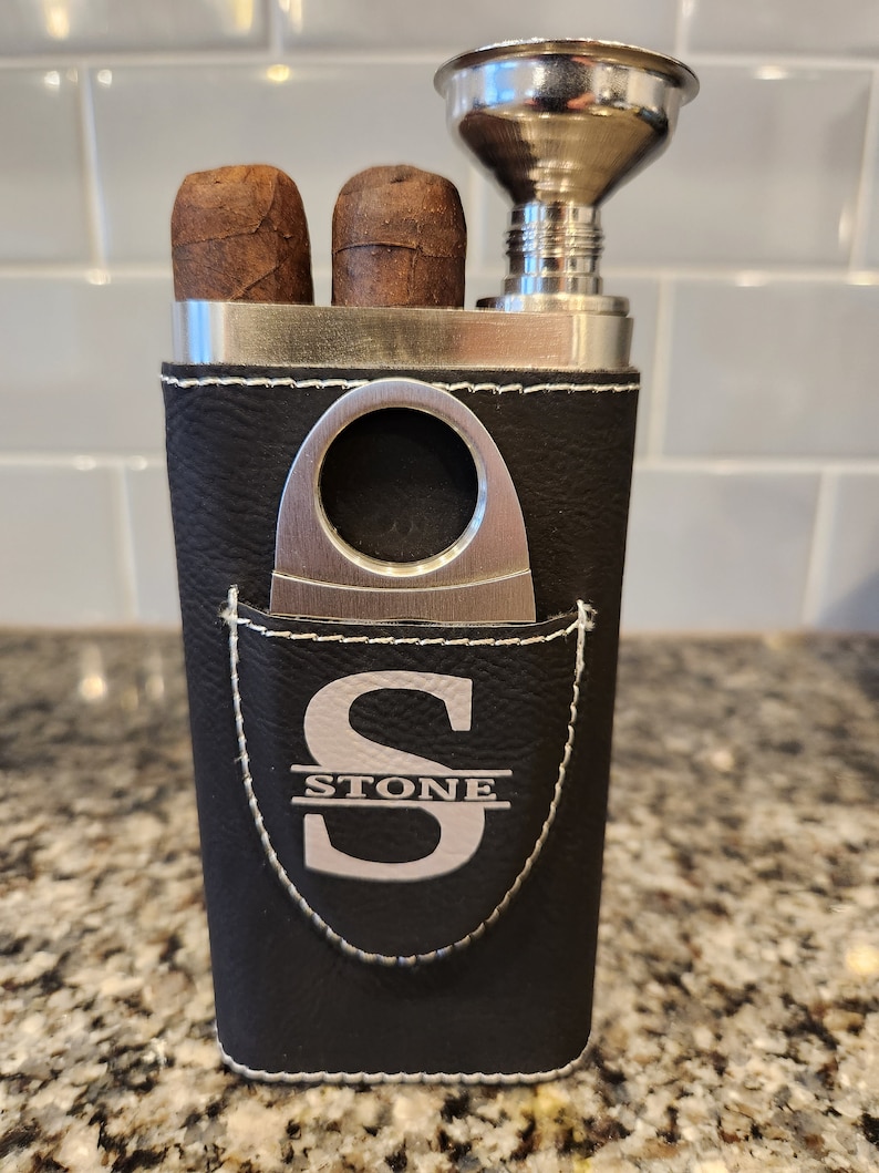 May include: A black leather cigar case with a silver metal cigar cutter, a silver metal funnel, and two cigars. The case has a pocket with a silver metal monogram that says "STONE".