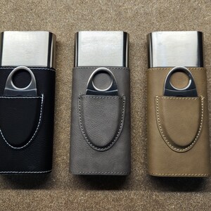 May include: Three leather cigar cases with metal cigar cutters. The cases are black, grey, and brown. Each case has a metal cutter inside a leather pocket.