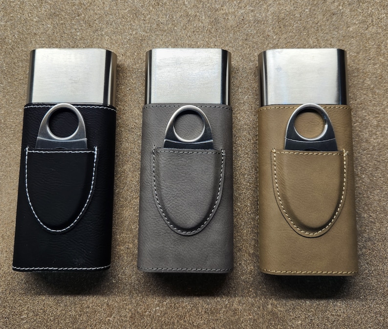 May include: Three leather cigar cases with metal cigar cutters. The cases are black, grey, and brown. Each case has a metal cutter inside a leather pocket.