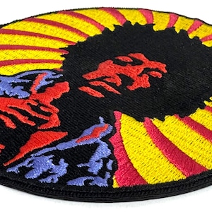 Può includere: Toppa ricamata con un design vibrante. La toppa presenta un centro nero con una figura rossa, circondata da raggi gialli e rossi. La parte inferiore presenta forme rosse e blu.