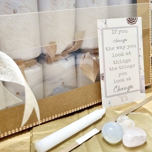 May include: A set of six white Christmas crackers in a clear plastic box with a brown cardboard base. The box is decorated with a gold glitter trim. The crackers are tied with brown ribbon and have a white paper label with gold lettering. The box also includes a white candle, a small piece of paper with the text "If you change the way you look at things the things you look at Change", and three small white stones.