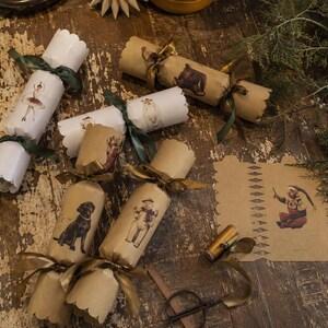 May include: A collection of Christmas crackers in white and kraft brown paper, tied with dark green and gold ribbons. The crackers feature illustrations of a ballerina, a dog, a snowman, and a small figure, with scalloped edges on the ends.