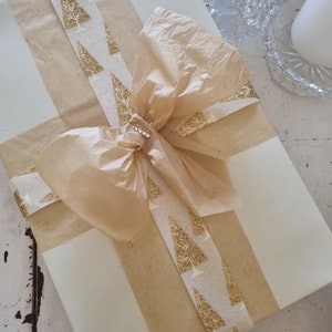 May include: A white gift box wrapped with brown paper and a gold ribbon with a pattern of glitter Christmas trees. A brown tissue paper bow is tied on top of the box.