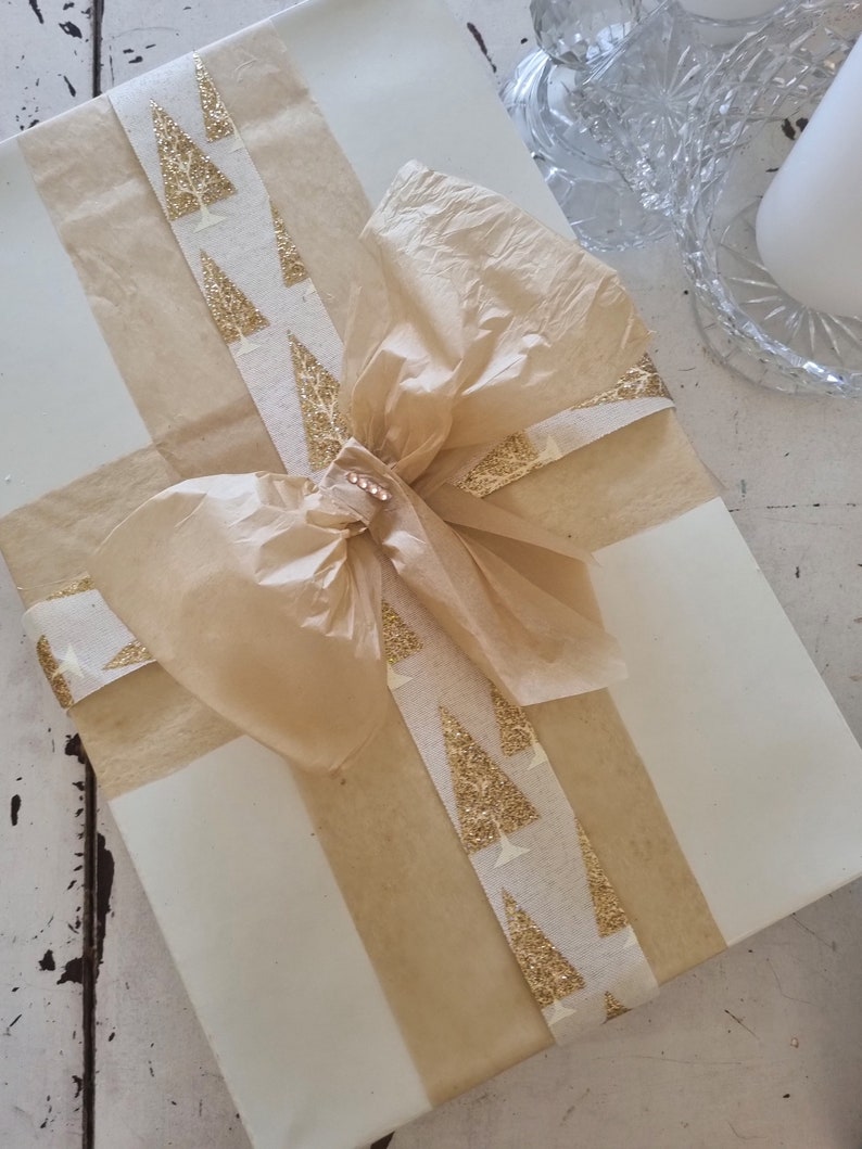 May include: A white gift box wrapped with brown paper and a gold ribbon with a pattern of glitter Christmas trees. A brown tissue paper bow is tied on top of the box.