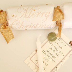 May include: A white Christmas cracker with gold ribbon and gold glitter. The cracker has the words "Merry Christmas" written on it. A small white candle, a green stone, and a card with the text "If you change the way you look at things, the things you look at change" are also in the image.