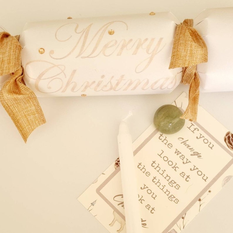 May include: A white Christmas cracker with gold ribbon and gold glitter. The cracker has the words "Merry Christmas" written on it. A small white candle, a green stone, and a card with the text "If you change the way you look at things, the things you look at change" are also in the image.