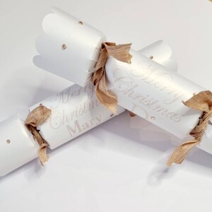 May include: Two white Christmas crackers with gold ribbon and gold glitter accents. The crackers are personalised with the text "Merry Christmas Mary" and "Merry Christmas Dad".