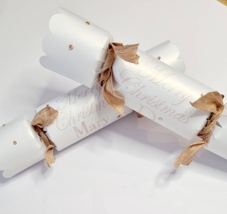 May include: Two white Christmas crackers with gold ribbon and gold glitter accents. The crackers are personalised with the text "Merry Christmas Mary" and "Merry Christmas Dad".